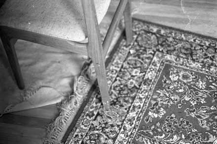a black and white photograph of a dirty wood floor and rugs and the legs of a midcentury modern dining chair