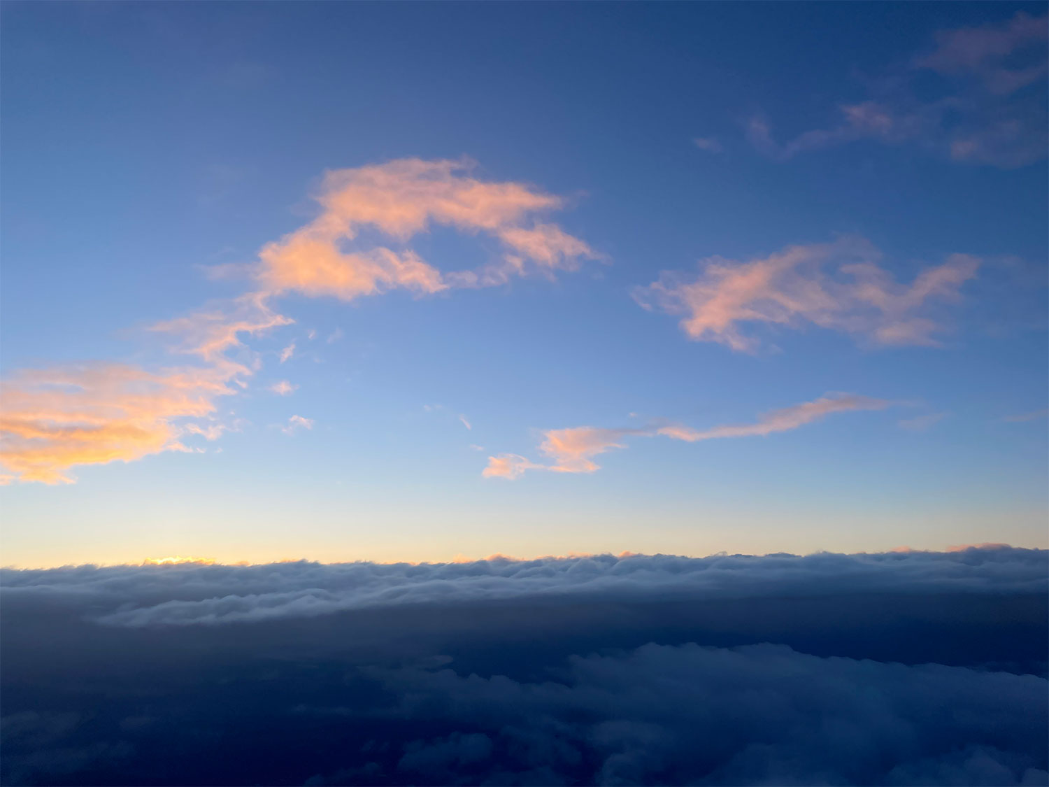 Looking east in the morning from above the clouds; that is, an image of the view out the window of an airplane, with a few wispy clouds pale pink above the gloaming