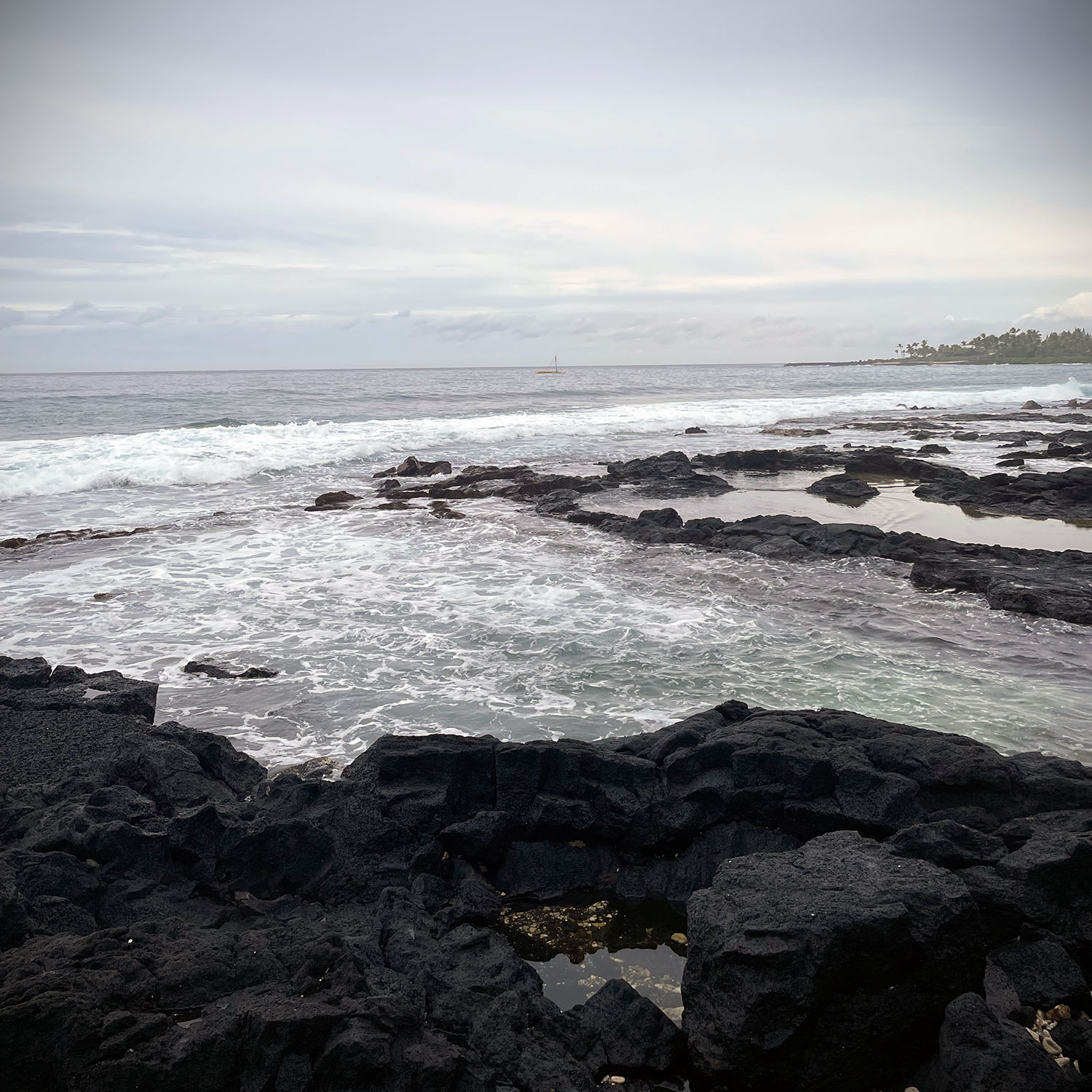 A view of the Pacific looking more or less west from Kailua Kona on a cloud day; black lava rocks in the foreground, blue water, and slate sky, with a narrow, blurred line of a land and palm trees in the distance.