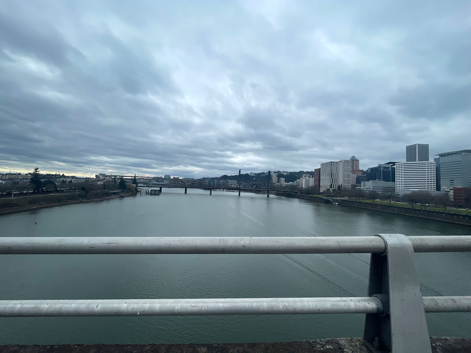 A view south from the Morrison Bridge in Portland, OR, under a cloudy sky, over wind-riffled water, to a distant urban skyline, partially blocked by the railing on the bridge