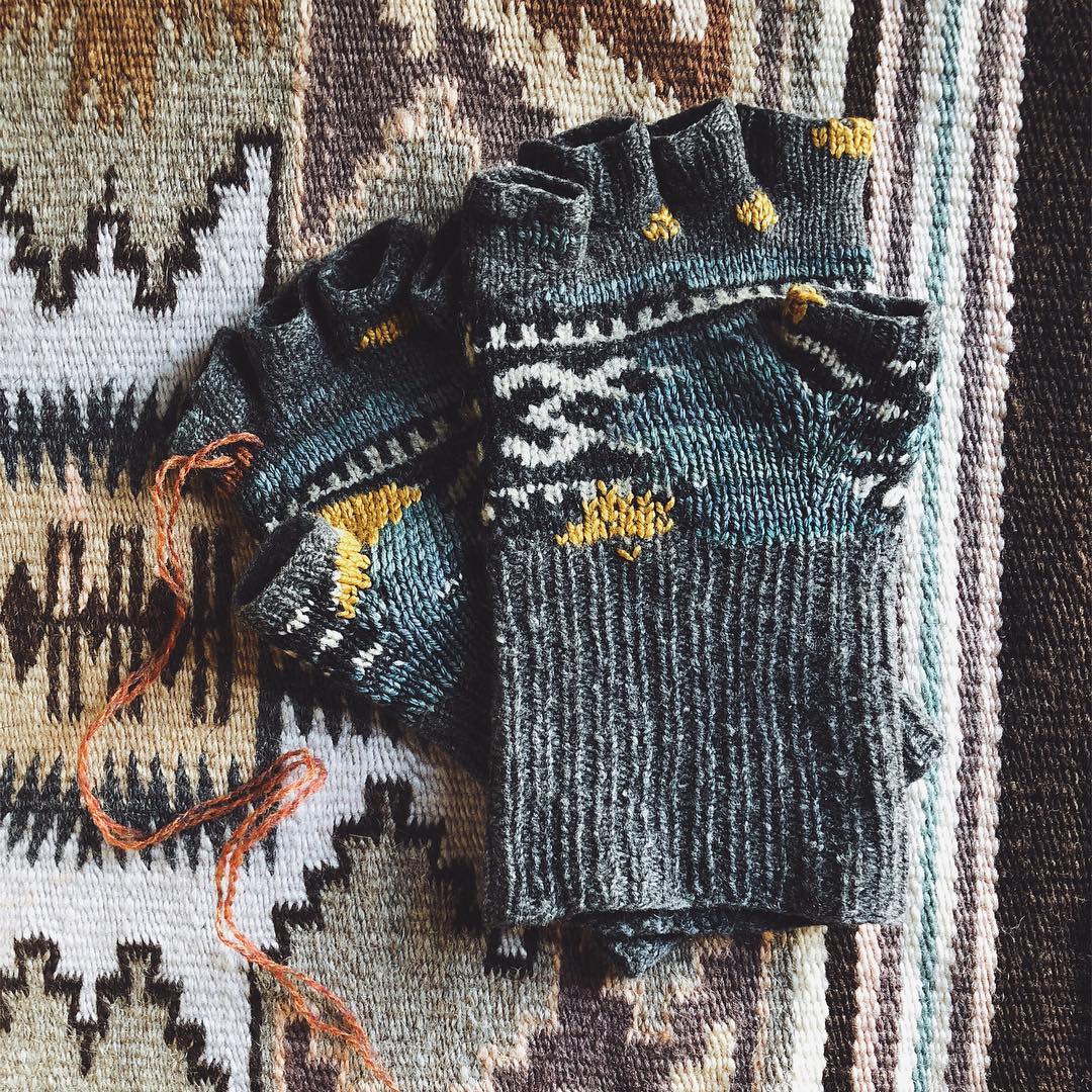 A pair of grey handknit colorwork fingerless gloves, with mending in diverse colors