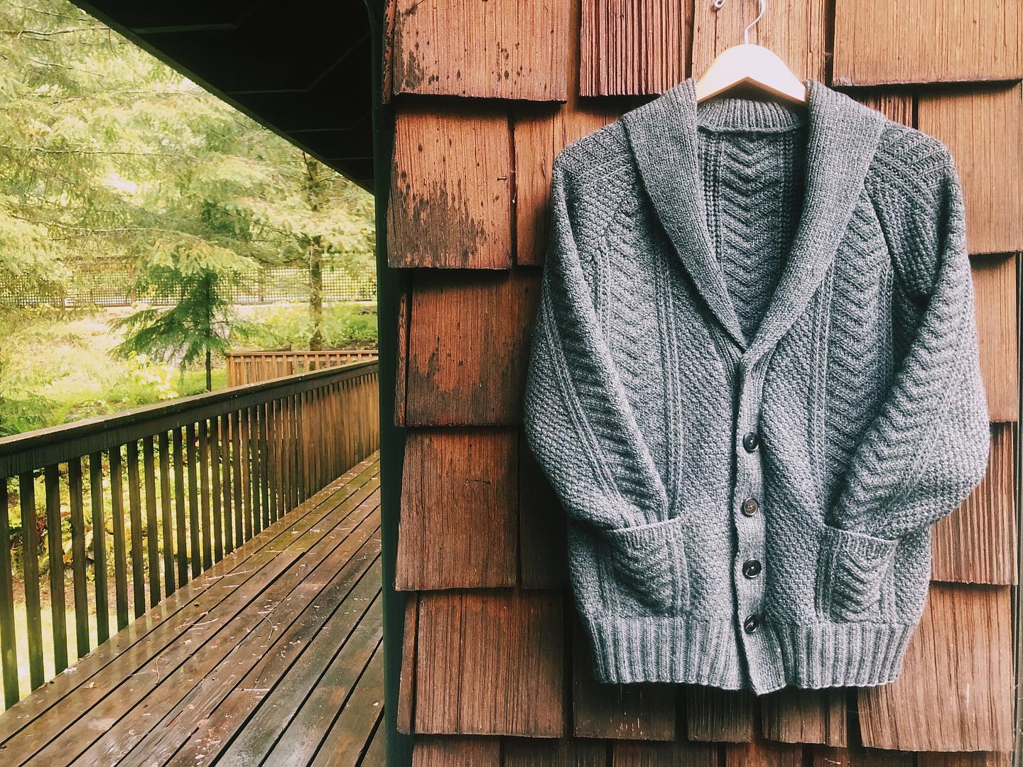 An image of a grey textured cardigan with a shawl collar and sleeves shoved into pockets, hung on a hanger against the cedar shakes on the corner of a cabin. The background is very green and springlike.